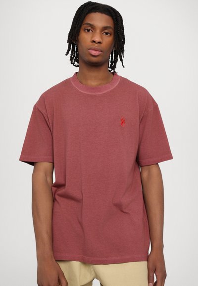 Camiseta de algodón color púrpura con mangas cortas, cuello redondo y logo de llama roja en el pecho; textura suave y corte relajado.