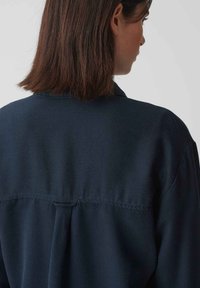 Navy blue button-up shirt featuring a collar, back pleat, and a slightly structured texture, with a visible yoke detail at the shoulders.