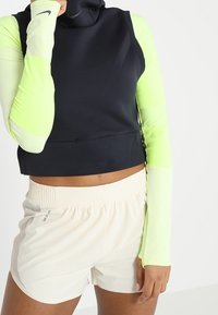 Czarny krótki strój sportowy z wysokim kołnierzem i długimi neonowozielonymi rękawami, zestawiony z jasnobeżowymi szortami z elastycznym pasem.