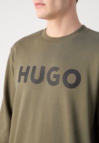 Svetle olivově zelená mikina s kulatým výstřihem a oversize střihem, s nápisem "HUGO" tištěným tučnými černými písmeny přes hruď.