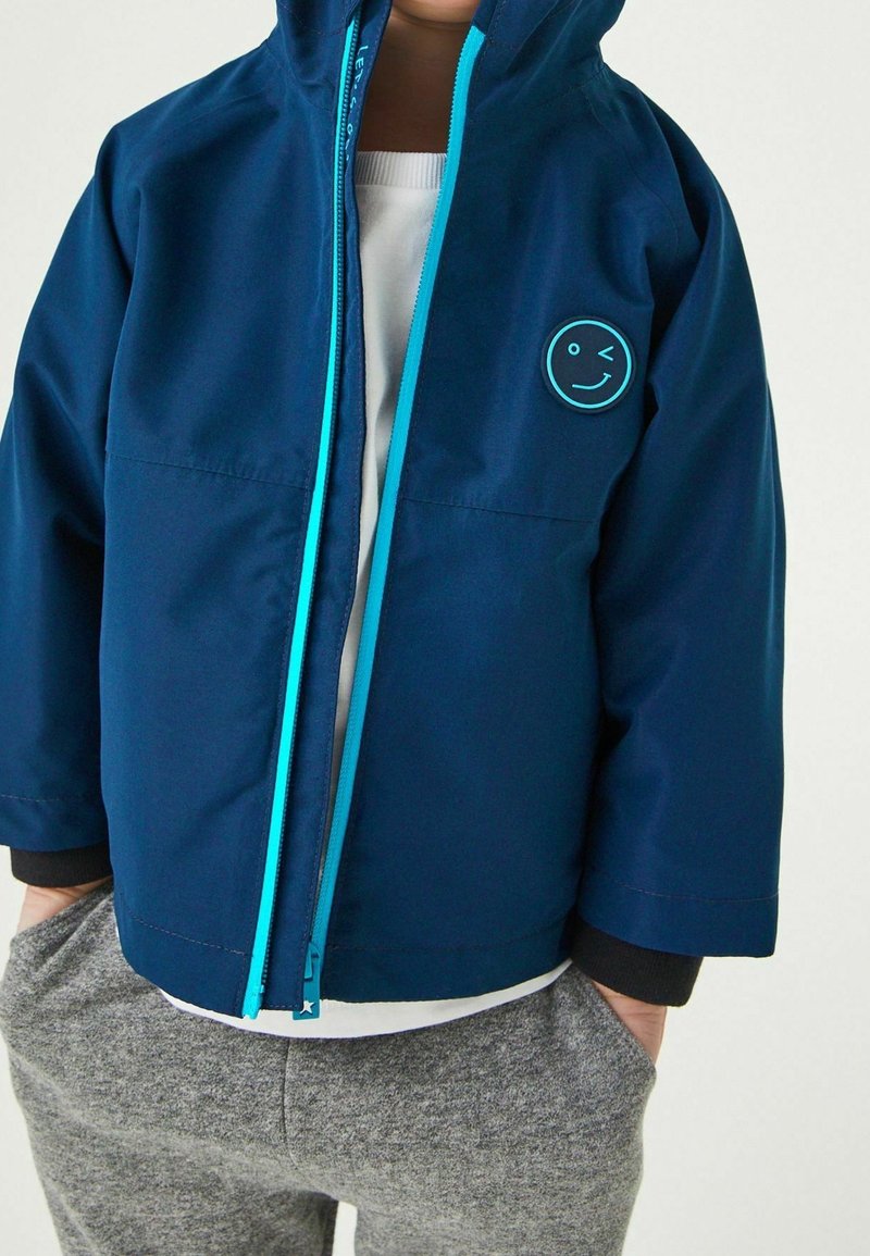 Enfant portant une veste à capuche bleue avec une fermeture éclair bleu clair et un patch smiley faisant un clin d’œil sur la poitrine gauche.