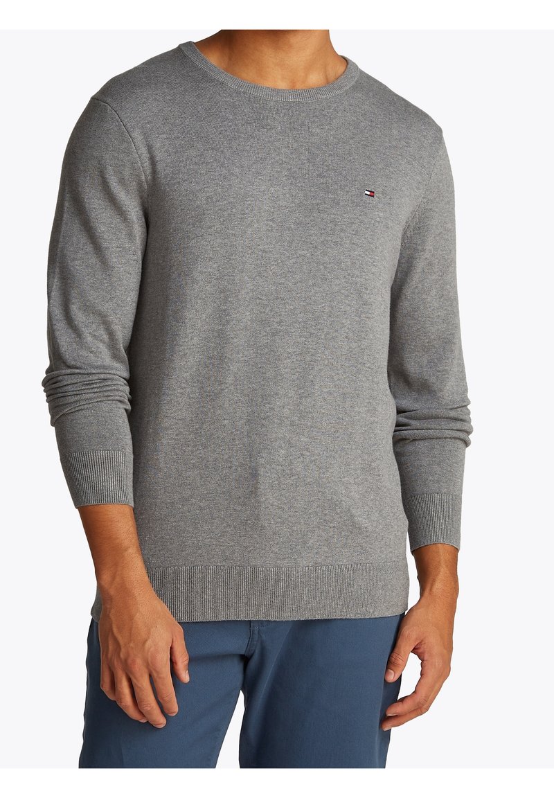 Homme portant un pull gris ajusté à col rond avec un petit logo sur la poitrine et un pantalon bleu, debout les bras détendus le long du corps.