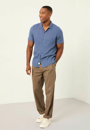 Kurzärmeliges, hellblaues Hemd mit einem strukturierten Streifenmuster, kombiniert mit hellbraunen, weit geschnittenen Hosen und weißen Turnschuhen, stehend vor einem neutralen Hintergrund.