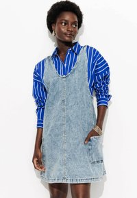 Robe salopette en denim délavé, dotée d'un décolleté en scoop, de deux poches latérales et d'une coupe décontractée, portée sur une chemise à rayures bleues avec fermeture à boutons.
