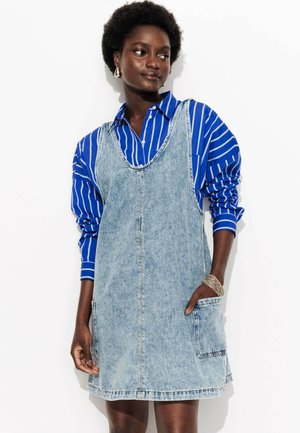 Robe salopette en denim délavé, dotée d'un décolleté en scoop, de deux poches latérales et d'une coupe décontractée, portée sur une chemise à rayures bleues avec fermeture à boutons.