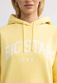 Geltonas pūkinis džemperis, pagamintas iš medvilnės, su baltu nusidėvėjusiu spaudiniu, kuris skaito "BIG STAR JEANS", su raišteliais ir laisvu siluetu.