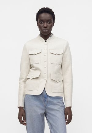 Personne à la peau foncée portant une veste blanche texturée à boutons avec quatre poches à rabat et un jean bleu clair, debout sur un fond blanc uni.