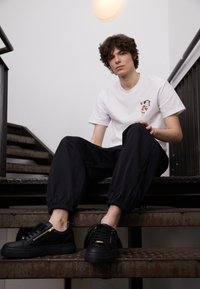 Baskets décontractées noires avec un détail zippé, associées à un pantalon avec bords élastiques noirs et une chemise blanche arborant un motif floral.