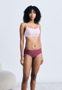 Γυναίκα με σγουρά μαλλιά που φοράει ροζ ανοιχτό μπραλέτ και σετ μωβ εσώρουχα χωρίς ραφές, στέκεται μπροστά σε απλό γκρι φόντο με μία μπεζ καρέκλα κοντά της.