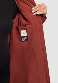 Rode, lichtgewicht jas met een soepele textuur. Binnenkant heeft een label en een zak. Geopend om details van de binnenvoering te tonen.