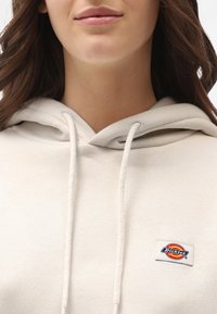 Felpa leggera color beige con cappuccio e cordino, presenta una toppa rettangolare con logo in rosso, giallo e blu con il testo "Dickies". Tessuto morbido.
