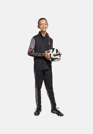 Schwarze Sportjacke mit grauen Ärmeln, roten Akzenten, passenden schwarzen Hosen und schwarzen Fußballschuhen. Das Model hält einen schwarz-weißen Fußball.