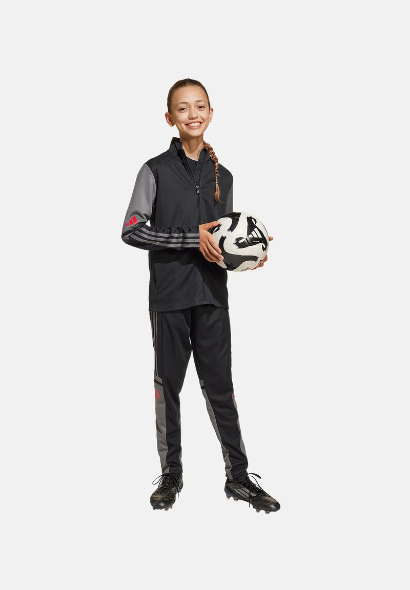 Schwarze Sportjacke mit grauen Ärmeln, roten Akzenten, passenden schwarzen Hosen und schwarzen Fußballschuhen. Das Model hält einen schwarz-weißen Fußball.