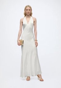 Γυναίκα που φοράει λευκό σατέν φόρεμα με λαιμόκοψη halter, κρατώντας χρυσό clutch με υφή, φορώντας χρυσά σανδάλια με τακούνι και βραχιόλι.