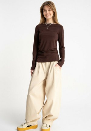 Młoda kobieta uśmiechająca się, ubrana w brązową bluzkę z długim rękawem, beżowe szerokie spodnie i beżowe trampki z żółtymi podeszwami, stojąca z rękami w kieszeniach.
