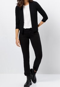 Schwarze, leichte Strickjacke mit offener Vorderseite und Dreiviertelärmeln, über einer weißen Bluse getragen. Kombiniert mit eng anliegenden schwarzen Jeans und Stiefeletten.