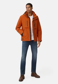Pomarańczowa wodoodporna kurtka z zamkiem błyskawicznym i zapięciami na napy, dwoma przednimi kieszeniami i kapturem, zestawiona z niebieskimi dżinsami i brązowymi butami.