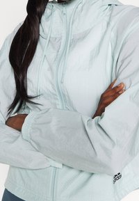 Person med korslagda armar, bär en ljusblå Adidas-zip-jacka med dragsnören och långt mörkt hår över ena axeln.