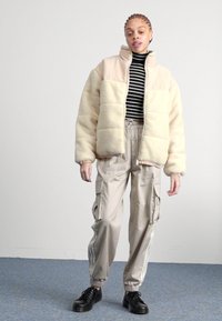 Beige plyschjacka med hög krage, svart och vit randig polotröja, ljusbeiga cargobyxor och svarta snörskor.
