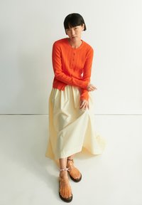 Oranger, gerippter Cardigan mit Knöpfen, über einem cremefarbenen Maxirock getragen, kombiniert mit transparenten Sandalen mit Riemen.
