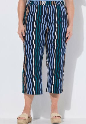 Frau trägt kurze Hose mit vertikal gewellten Streifen in Blau, Grün, Weiß und Braun, kombiniert mit beigen, gewebten Plateau-Sandalen.