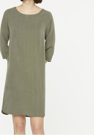 Žena nosící volné olivově zelené šaty délky ke kolenům s tříčtvrtečními rukávy a jednoduchým stříbrným náhrdelníkem.