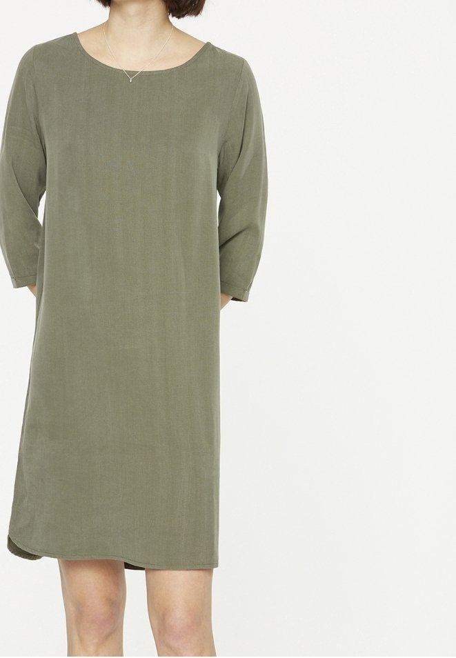 Femme portant une robe ample vert olive, longueur genou, avec des manches trois-quarts et un simple collier en argent.