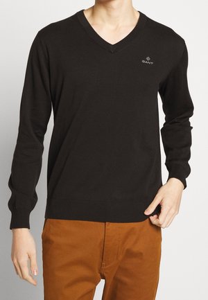 Homme portant un pull à col en V noir avec un petit logo GANT sur la poitrine et un pantalon marron, debout devant un fond uni.