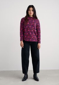 Langärmliger, fuchsiafarbener Pullover mit einem Blättermuster, kombiniert mit schwarzen, weit geschnittenen Hosen und schwarzen Ankle Boots. Glattes Material mit lockerer Passform.