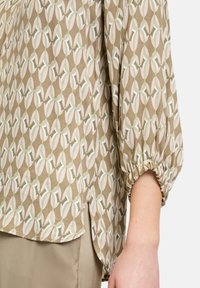 Blusa leggera beige con un motivo floreale geometrico. Presenta maniche a sbuffo con polsini elastici e un orlo leggermente curvato.
