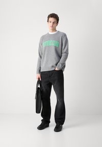 Grå genser med "FUTURE CLUB" i lys grønn, kombinert med svarte jeans og svarte sneakers, holder en svart veske. Jevn tekstur, avslappet passform.