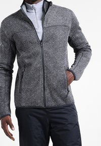 Giacca grigia con texture, collo alto e chiusura con zip, dotata di due tasche laterali e uno strato interno grigio chiaro. Abbinata a pantaloni scuri.