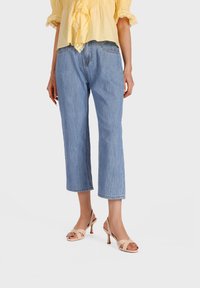 Mujer con vaqueros cropped azul claro, blusa amarilla y sandalias de tacón beige, de pie frente a un fondo liso.