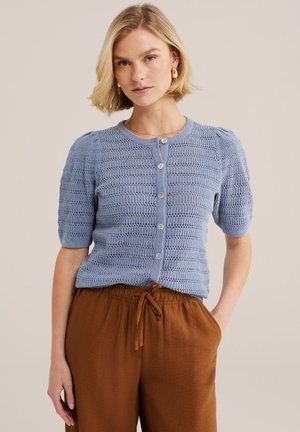 Kobieta ubrana w jasnoniebieski krótki sweter na guziki i brązowe spodnie ze sznurkiem, stoi z jedną ręką w kieszeni.