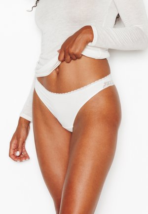 Donna che indossa biancheria intima bianca rifinita in pizzo e una maglia a maniche lunghe che solleva l'orlo per mostrare la biancheria intima su uno sfondo bianco.
