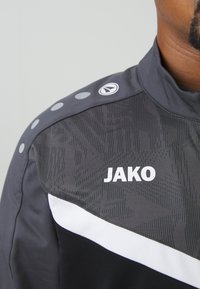 Grijze sportjack met een hoge kraag, textuurpatroon, witte streep en 'JAKO' logo in vetgedrukte witte tekst, met drukknoopaccenten op de schouder.