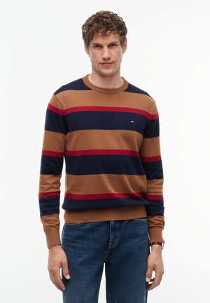 Homme portant un pull rayé marron, bleu marine et rouge et un jean bleu, debout devant un fond blanc uni.