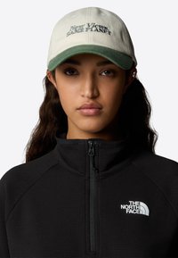 Gorra de algodón con visera verde y tela caqui, con texto bordado "Nuevas Vistas. MISMA TIERRA." Chaqueta de forro polar con cremallera negra con logo.