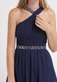 Mörkblå, enaxlad klänning med samlad textur, utsmyckad midja med metalliska detaljer och en plisserad kropp.