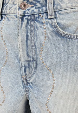 Lichtblauwe spijkerbroek close-up met koperen knoop en golvend patroon van kleine metalen studs langs voorzakken en naden.