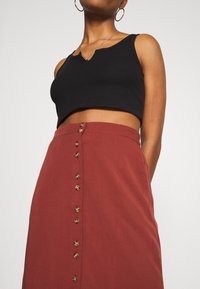 Femme portant un haut noir court sans manches et une jupe taille haute couleur rouille avec une rangée de boutons sur le devant, fond blanc.