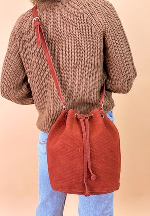 Roter Wildleder-Eimerbeutel mit einem Zugverschluss und Chevron-Muster, ausgestattet mit einem langen verstellbaren Tragegurt und weicher Textur.