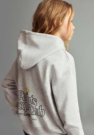 Junge Person mit welligem, blondem Haar, die einen hellgrauen Hoodie mit einem "Peanuts Snoopy Club"-Grafik auf dem Rücken trägt und seitlich schaut.