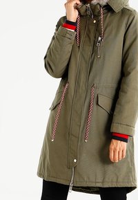 Parka verde olivo con cremallera, dos bolsillos frontales y cordones rojos y blancos. Cuenta con capucha y puños acanalados con un acento de color.