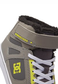 Zapatilla alta gris con forro de piel negra, logo amarillo, cordones blancos y textura con patrones. Presenta una correa de Velcro para un ajuste seguro.