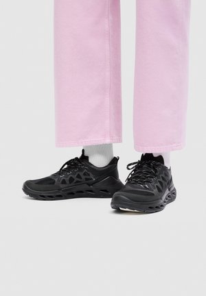 Persoon die roze broek en witte sokken draagt met zwarte sportschoenen met een patroon aan de zijkant en dikke zolen.