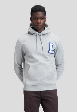 Uomo che indossa una felpa con cappuccio grigio chiaro con grandi lettere blu "L" e "LEE" sul petto, in piedi con le mani dietro la schiena su sfondo semplice.