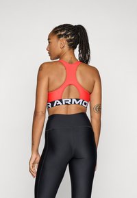 Sujetador deportivo rojo con diseño de espalda cruzada, que presenta un corte en la parte posterior y una banda negra ancha con texto blanco. Combinado con leggins negros.