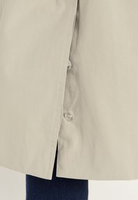 Beige coat hem with two clear buttons partially open, showing dark blue jeans underneath.