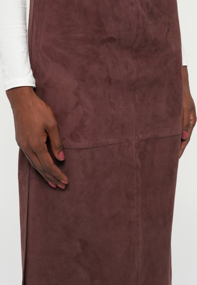 Vinrdeča semiš obleka z gladko teksturo in prilegajočo zasnovo, s šivnim detajlom v predelu pasu. Roka nežno počiva ob strani.